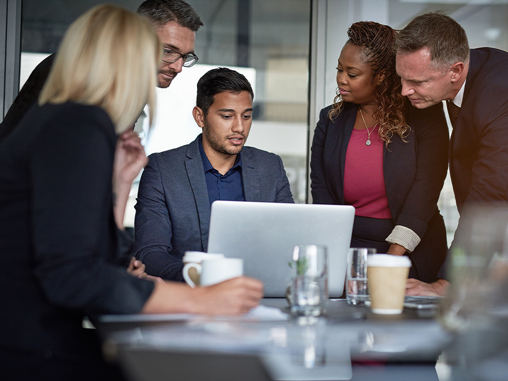 A group of diverse employees sitting around an apprentice's laptop in smart attire